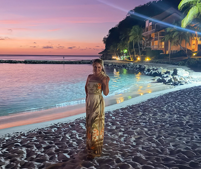 Cheryl on a Caribbean beach at dusk, a lit resort and palms behind her