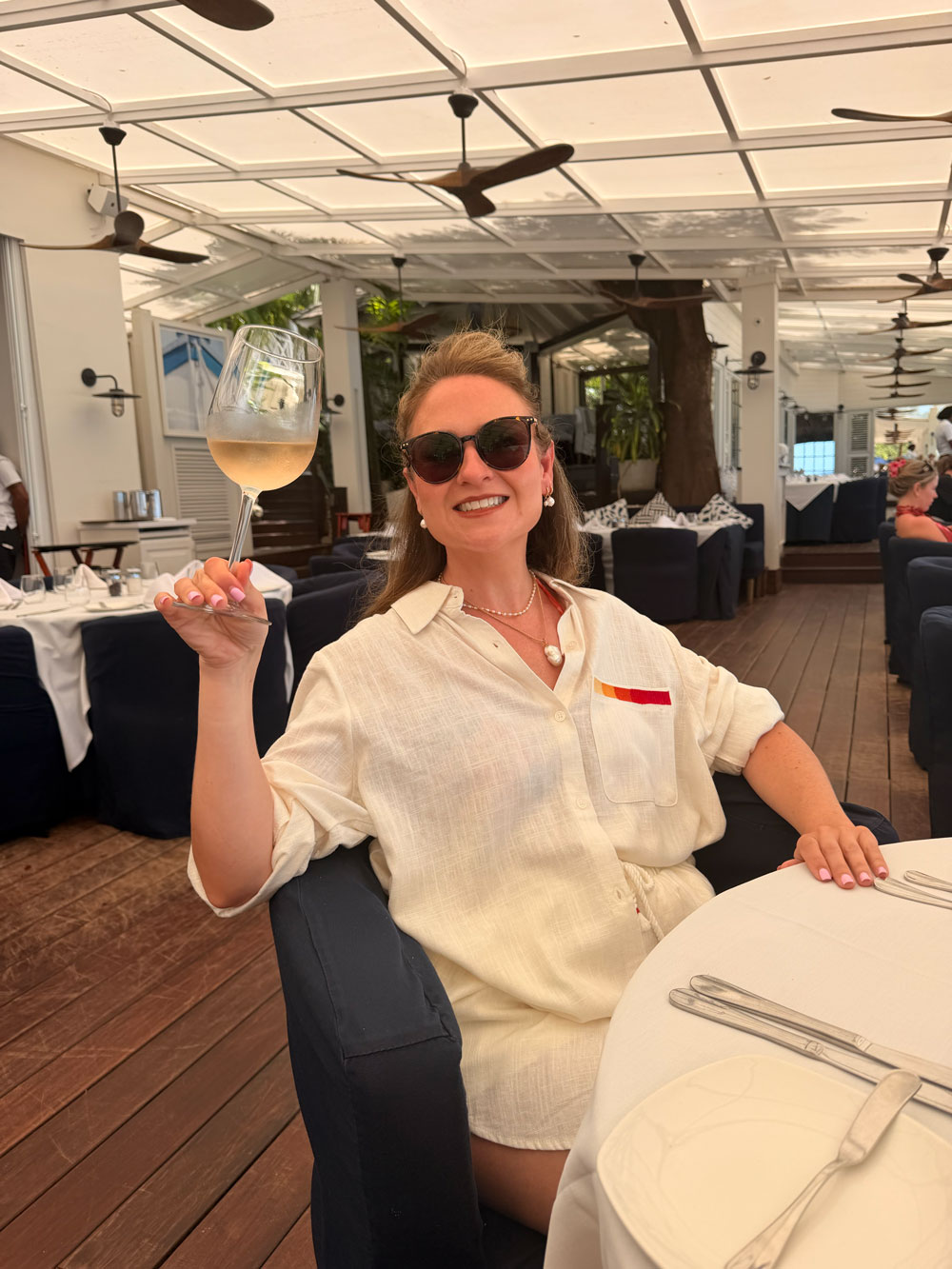 Cheryl raising a glass of chilled white wine at a laid table under
                    the ceiling fans at Lone Star, Barbados
