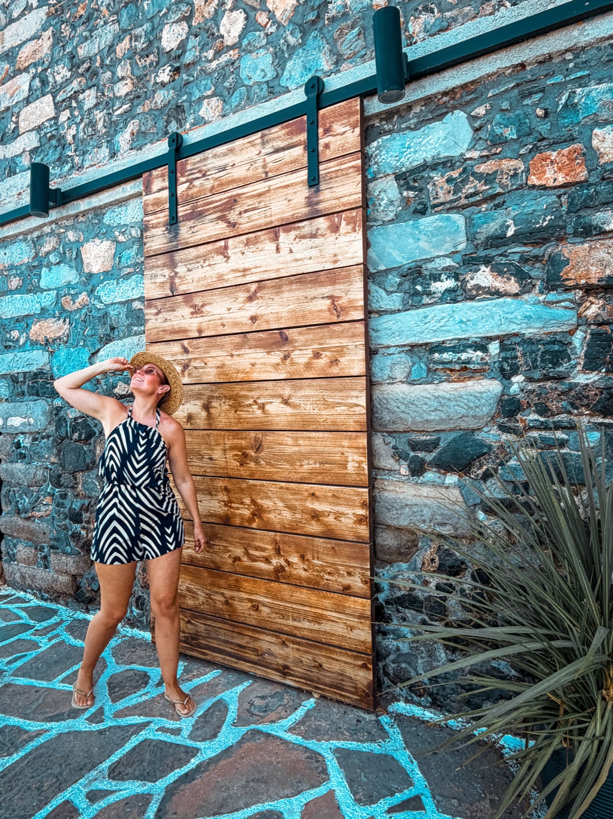 Cheryl in a straw hat beside a barn door set into a turquoise-washed
                    stone wall at a boutique hotel