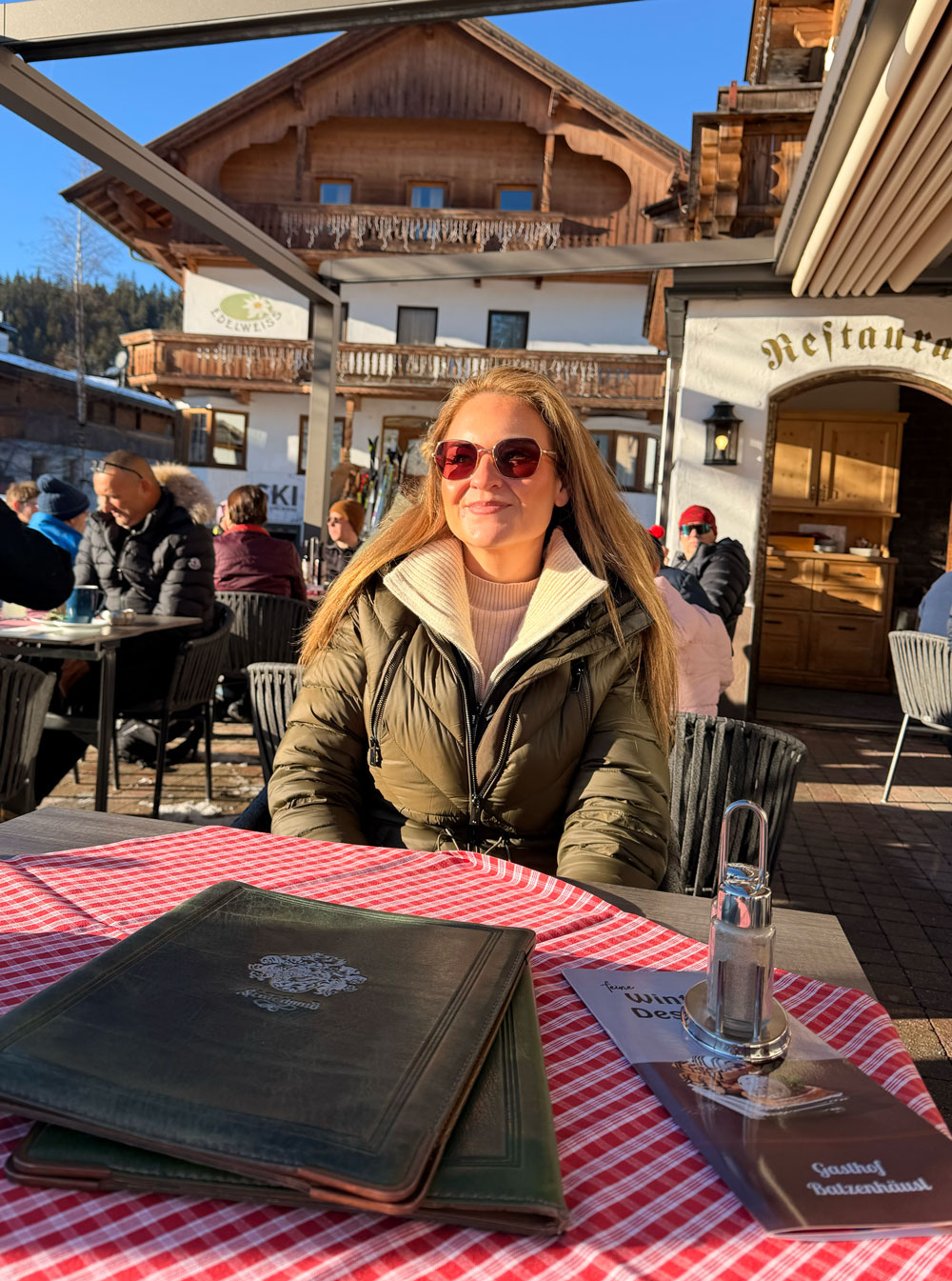 Cheryl at an outdoor restaurant table in the Austrian Alps, menus and a
                    red gingham cloth in front of her, a wooden chalet behind