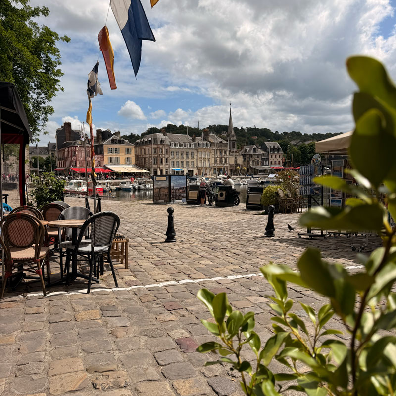 Cobbles, café chairs and flags on the harbour front at Honfleur, the
                    painted townhouses reflected in the water beyond