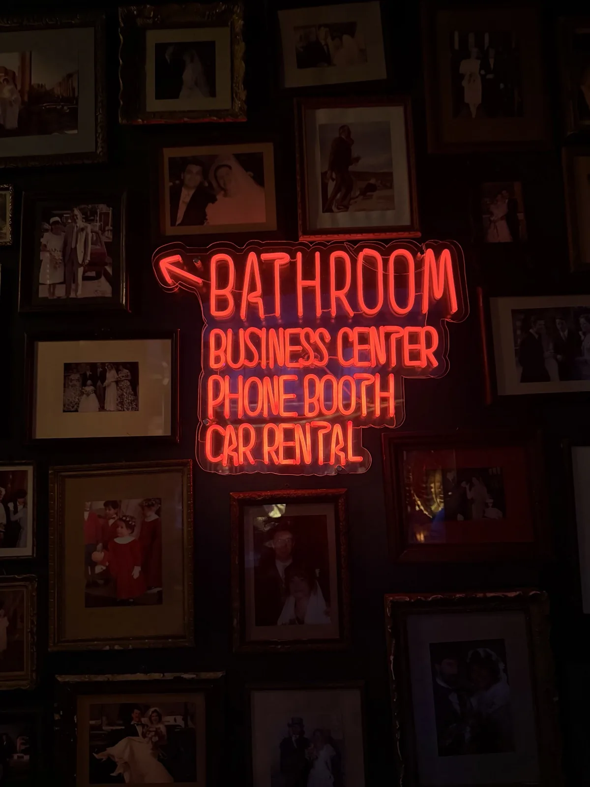 A red neon sign reading Bathroom, Business Center, Phone Booth, Car Rental above a wall of framed photographs