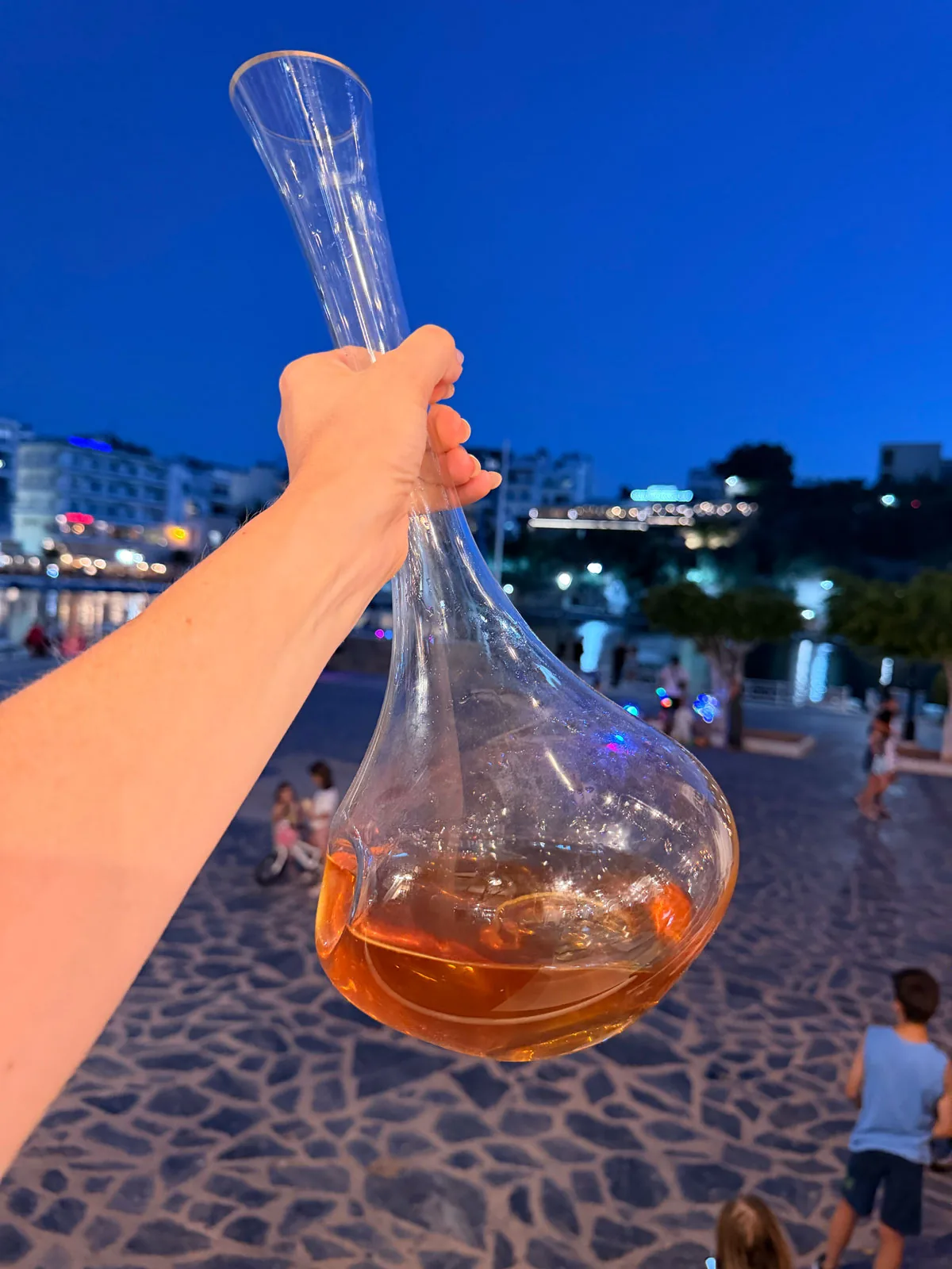 The Blanc des Coteaux held up in its glass decanter against the evening sky over Lake Voulismeni, nearly orange in the light