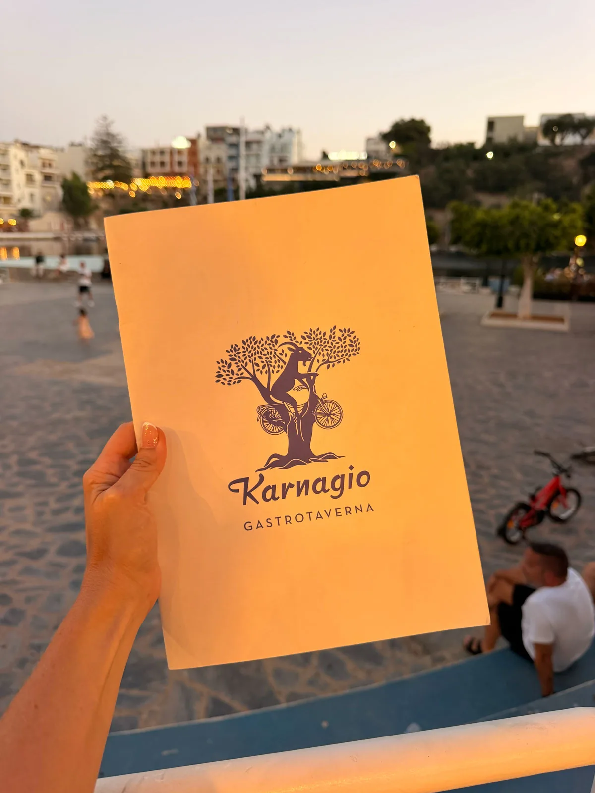 The Karnagio Gastrotaverna menu cover held up in front of Lake Voulismeni at dusk