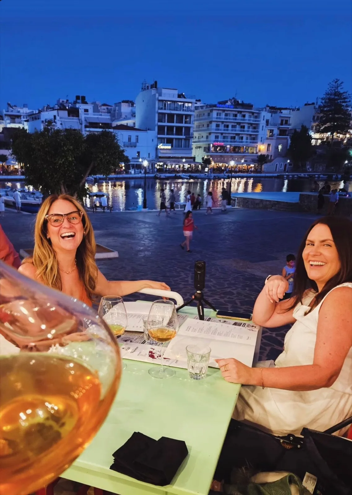 Laughing over wine at a green table at Karnagio, with Lake Voulismeni and the lights of Agios Nikolaos glowing behind