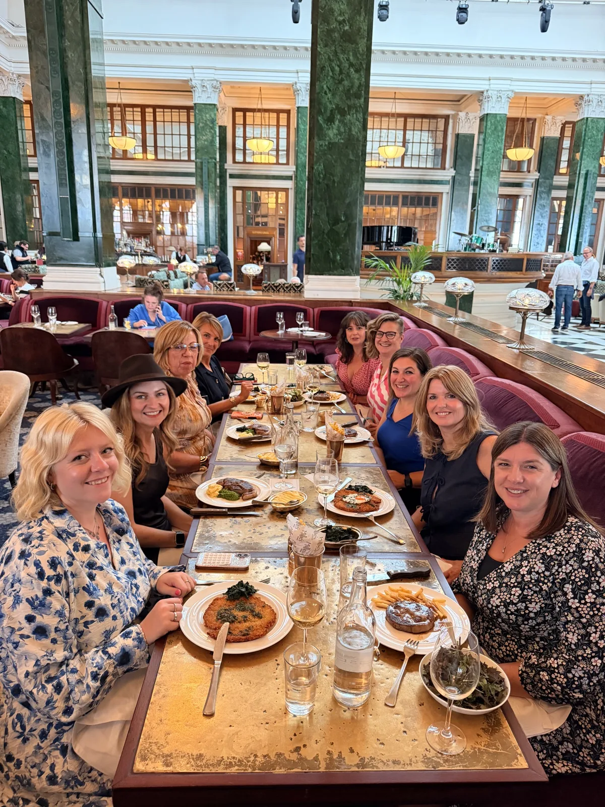 Nine of us at a long gold leaf table in the ground floor hall of The Ned, green marble columns and red leather banquettes behind