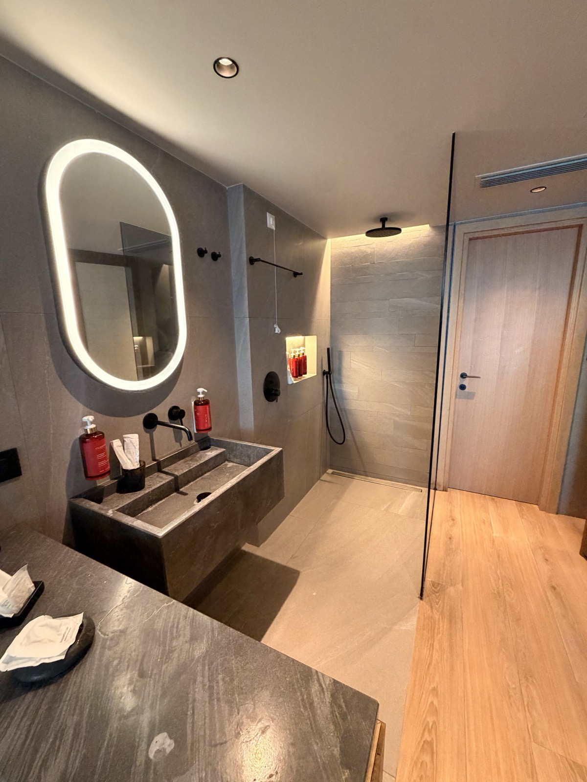 The open plan bathroom at NIKO Seaside Resort, with a backlit oval mirror, dark stone double basin and a walk in rain shower