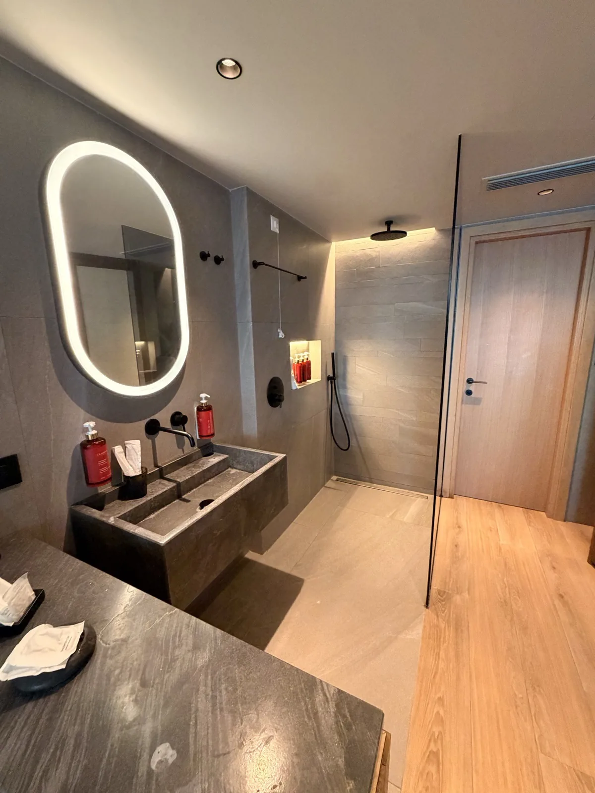 The open plan bathroom at NIKO Seaside Resort, with a backlit oval mirror, dark stone double basin and a walk in rain shower