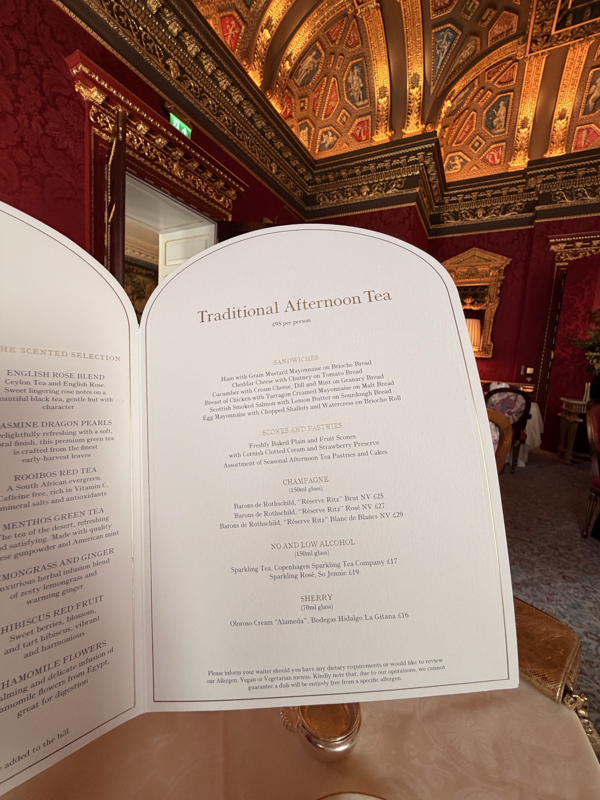 The afternoon tea menu at The Ritz London in front of crimson damask walls, gold framed mirrors and the ornate ceiling of William Kent House