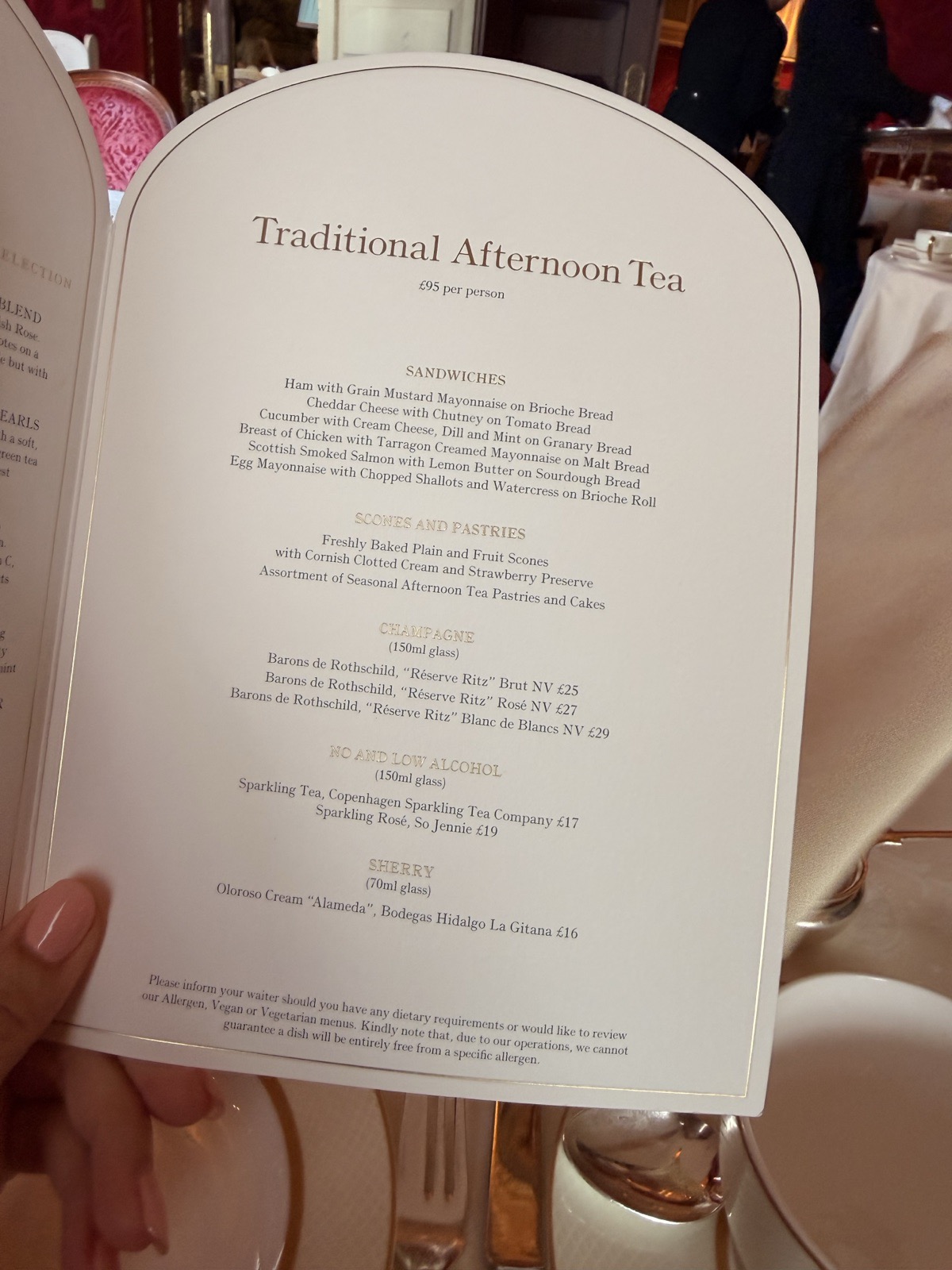 The Traditional Afternoon Tea menu at The Ritz London listing finger sandwiches, scones and champagne, held up in the red tea room